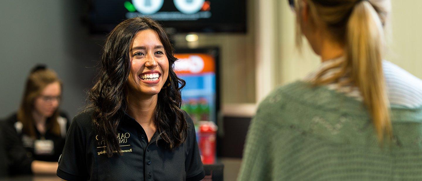 An employee at the front desk of the recreation center, speaking with a member.