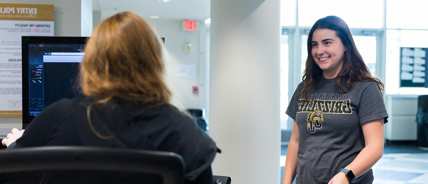 A woman wearing a Grizzlies shirt speaking to a person seated behind a desk.